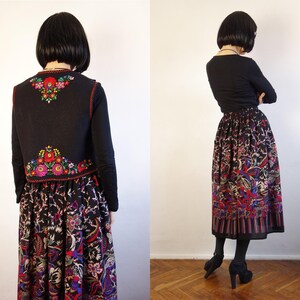 May include: A black waistcoat featuring vibrant floral embroidery in red, green, and yellow on the back, paired with a long, pleated skirt displaying a multi-coloured botanical print on a black background, worn over a plain black long-sleeve shirt.