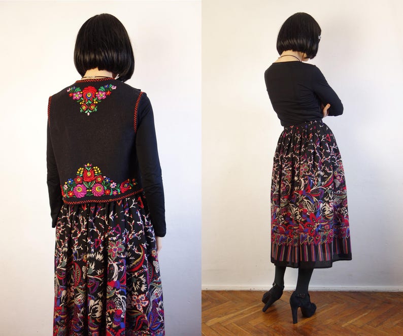 May include: A black waistcoat featuring vibrant floral embroidery in red, green, and yellow on the back, paired with a long, pleated skirt displaying a multi-coloured botanical print on a black background, worn over a plain black long-sleeve shirt.