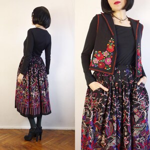 May include: A black long-sleeved top, a black waistcoat with red and green floral embroidery, and a black midi skirt featuring a vibrant, multi-coloured floral and paisley print. The skirt has a pleated waistline and side pockets, paired with black tights.