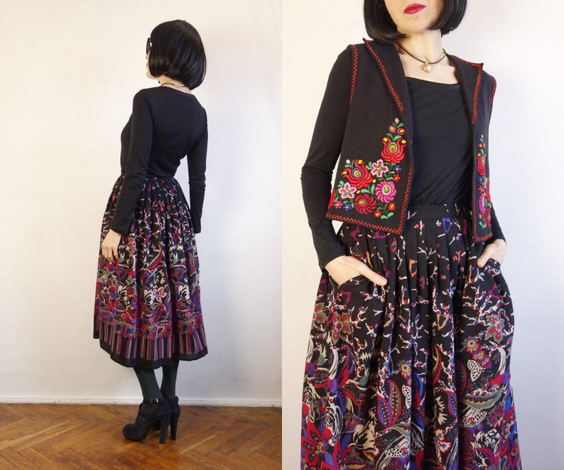 May include: A black long-sleeved top, a black waistcoat with red and green floral embroidery, and a black midi skirt featuring a vibrant, multi-coloured floral and paisley print. The skirt has a pleated waistline and side pockets, paired with black tights.