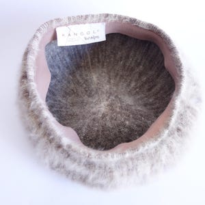 May include: A light gray Kangol design beret with a soft, fuzzy texture. The beret has a pink interior band and a tag with the Kangol logo. The hat is a classic style, suitable for various occasions.