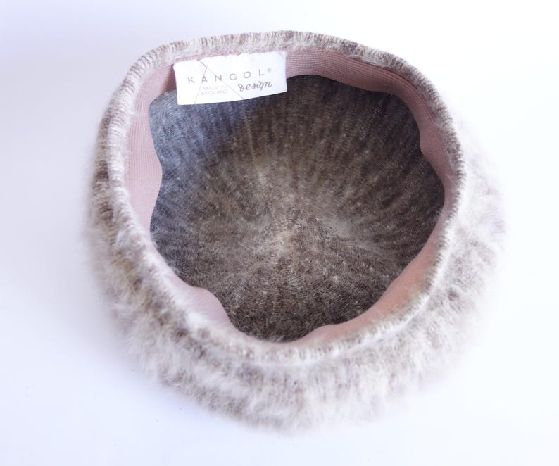 May include: A light gray Kangol design beret with a soft, fuzzy texture. The beret has a pink interior band and a tag with the Kangol logo. The hat is a classic style, suitable for various occasions.