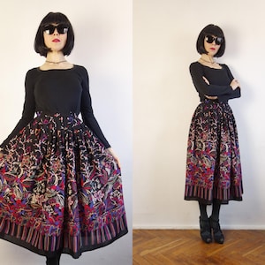 Puede incluir: Una falda larga plisada con base negra y un estampado floral y de cachemira multicolor vibrante. El dobladillo presenta un borde de rayas verticales en tonos rojo, azul y morado. La falda se combina con una camiseta negra de manga larga.