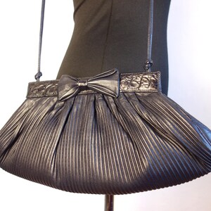 May include: A metallic gray pleated handbag with a bow detail. The bag has a long, thin strap and a textured top panel. The bow is centered on the front, and the pleats create a fan-like shape. The bag is displayed on a mannequin.