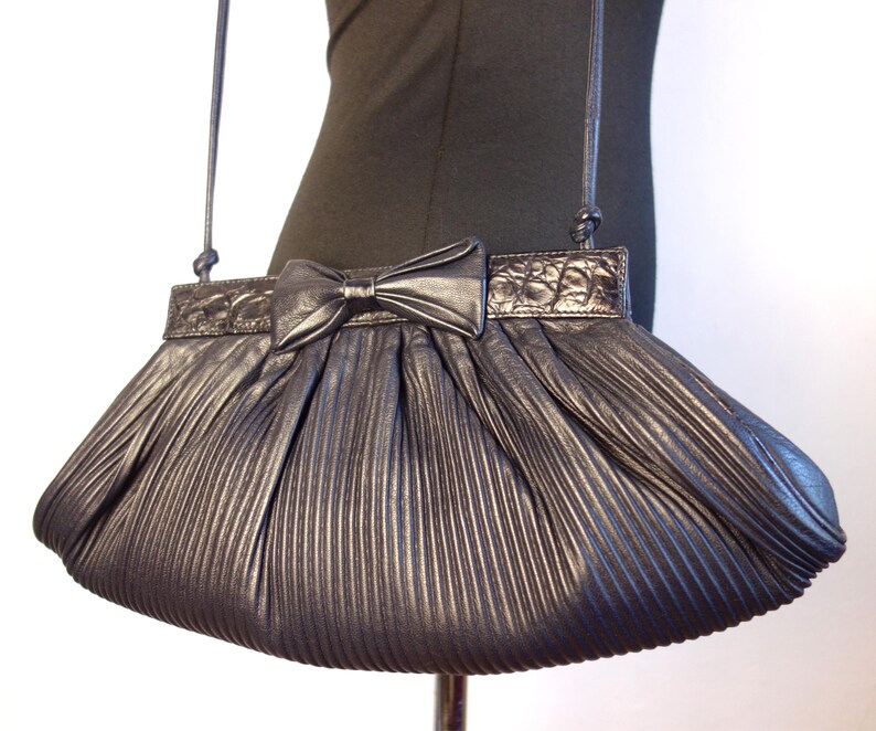 May include: A metallic gray pleated handbag with a bow detail. The bag has a long, thin strap and a textured top panel. The bow is centered on the front, and the pleats create a fan-like shape. The bag is displayed on a mannequin.