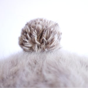 May include: Close-up of a light brown and white pom-pom on top of a light brown, fuzzy surface. The pom-pom is round and fluffy, with a mix of light and dark brown fibers. The fuzzy surface is soft and textured.