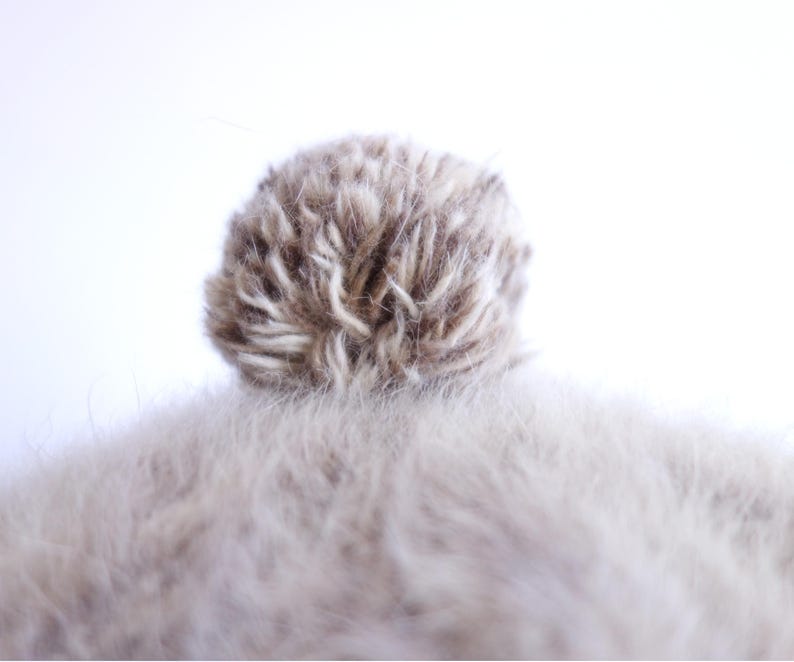 May include: Close-up of a light brown and white pom-pom on top of a light brown, fuzzy surface. The pom-pom is round and fluffy, with a mix of light and dark brown fibers. The fuzzy surface is soft and textured.