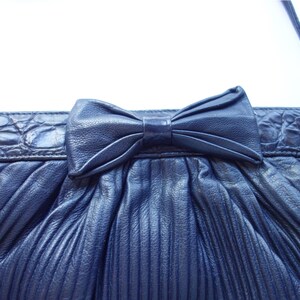 May include: Navy blue pleated clutch purse with a bow detail. The purse features a textured, crocodile-like trim and a thin shoulder strap. The bag is made of a soft, pleated material.