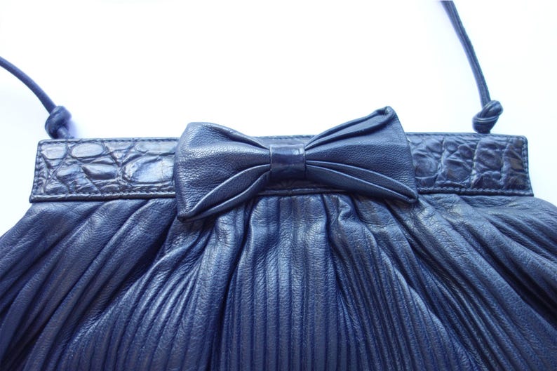 May include: Navy blue pleated clutch purse with a bow detail. The purse features a textured, crocodile-like trim and a thin shoulder strap. The bag is made of a soft, pleated material.