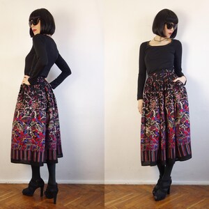 May include: A long, pleated skirt featuring a dark base with a vibrant, multi-coloured floral and abstract pattern, paired with a solid black long-sleeved top. The skirt has a wide, striped border at the hem and a full, flowing silhouette.