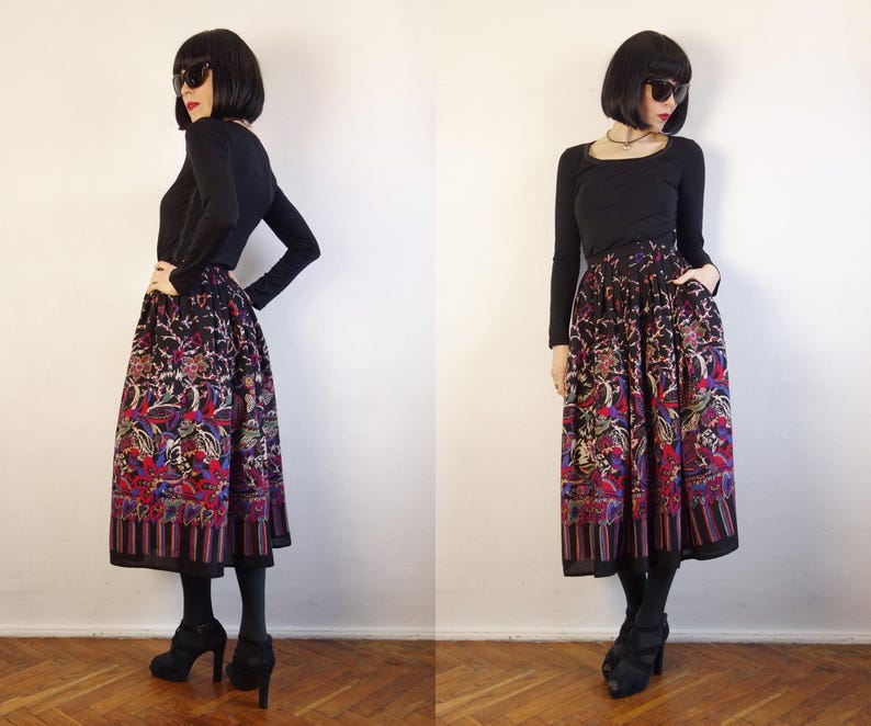 May include: A long, pleated skirt featuring a dark base with a vibrant, multi-coloured floral and abstract pattern, paired with a solid black long-sleeved top. The skirt has a wide, striped border at the hem and a full, flowing silhouette.