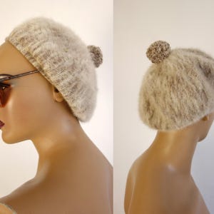 May include: A beige beret with a fluffy texture and a small pom-pom on top. The hat is shown on a mannequin head, with the side profile visible. The beret is a neutral color, suitable for various outfits.