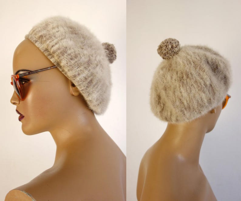 May include: A beige beret with a fluffy texture and a small pom-pom on top. The hat is shown on a mannequin head, with the side profile visible. The beret is a neutral color, suitable for various outfits.