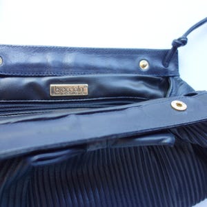 May include: Navy blue leather handbag with a pleated design. The interior features a zipper pocket and a gold-toned brand label. The bag has a long, thin strap and gold-colored snap closures.