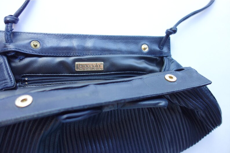 May include: Navy blue leather handbag with a pleated design. The interior features a zipper pocket and a gold-toned brand label. The bag has a long, thin strap and gold-colored snap closures.