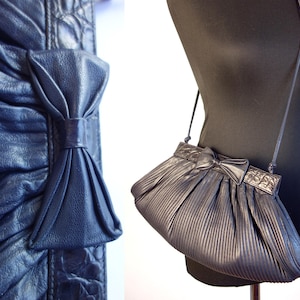 May include: A dark blue, pleated shoulder bag with a bow detail. The bag has a long, thin strap and a rectangular top with a decorative pattern. The bag is displayed on a black mannequin.