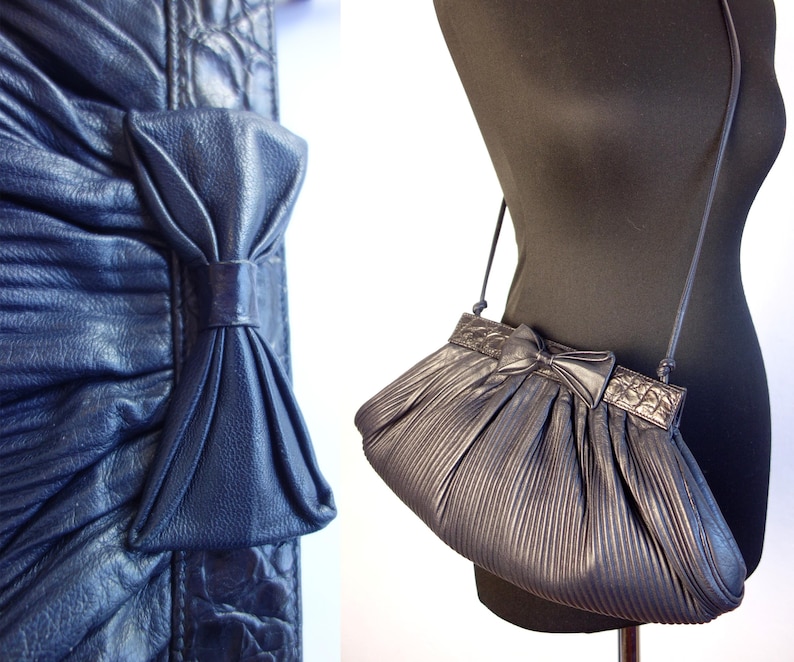 May include: A dark blue, pleated shoulder bag with a bow detail. The bag has a long, thin strap and a rectangular top with a decorative pattern. The bag is displayed on a black mannequin.