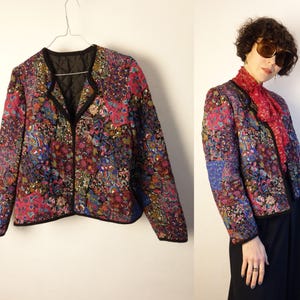 Op de afbeelding: Een gewatteerde jas met een levendig bloemenpatroon in roze, blauwe en groene tinten. De jas heeft een zwarte bies en een V-hals. De jas wordt op een hanger getoond en door een persoon gedragen.