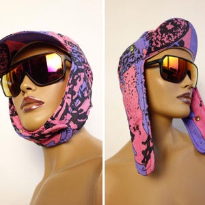 Pode incluir: Um gorro acolchoado com abas para as orelhas com um padrão abstrato vibrante de salpicos em rosa, roxo e preto, combinado com óculos de sol grandes com armações escuras e lentes refletoras de tom laranja, exibido a partir de dois ângulos.