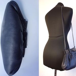 May include: A navy blue, pleated leather handbag with a long strap, displayed on a black mannequin. The bag has a rounded shape with a gathered top and a small bow detail. The leather has a textured, ribbed appearance.
