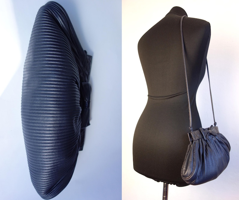 May include: A navy blue, pleated leather handbag with a long strap, displayed on a black mannequin. The bag has a rounded shape with a gathered top and a small bow detail. The leather has a textured, ribbed appearance.