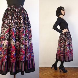 May include: A black midi skirt featuring a vibrant, multi-coloured floral and paisley pattern. The skirt has a high-waisted, pleated design that falls to mid-calf length, with a distinct vertical striped border along the bottom hemline.