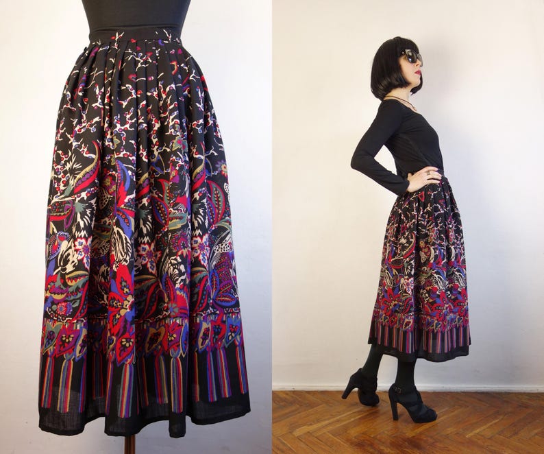 May include: A black midi skirt featuring a vibrant, multi-coloured floral and paisley pattern. The skirt has a high-waisted, pleated design that falls to mid-calf length, with a distinct vertical striped border along the bottom hemline.