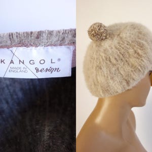 May include: A beige, fuzzy Kangol design hat with a pom-pom on top. The hat is paired with orange-tinted sunglasses. The label reads "KANGOL design MADE IN ENGLAND".