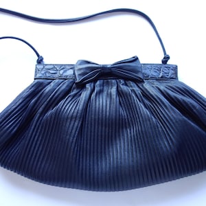 May include: A navy blue pleated handbag with a bow detail. The bag has a long, thin strap and a textured band across the top. The bag's design is a classic, elegant style.