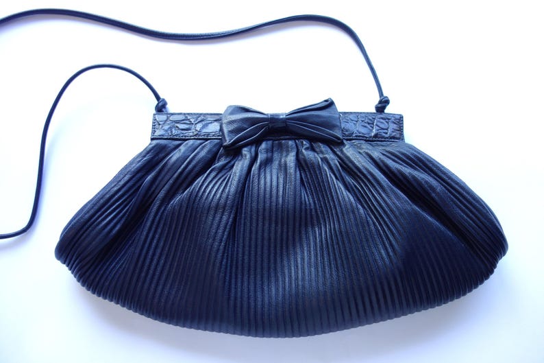 May include: A navy blue pleated handbag with a bow detail. The bag has a long, thin strap and a textured band across the top. The bag's design is a classic, elegant style.