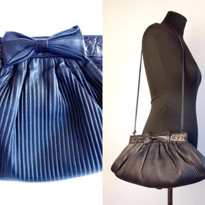 May include: A navy blue pleated handbag with a bow detail. The bag has a long strap and a textured top. The bag is displayed on a mannequin. The bag is a dark blue color.