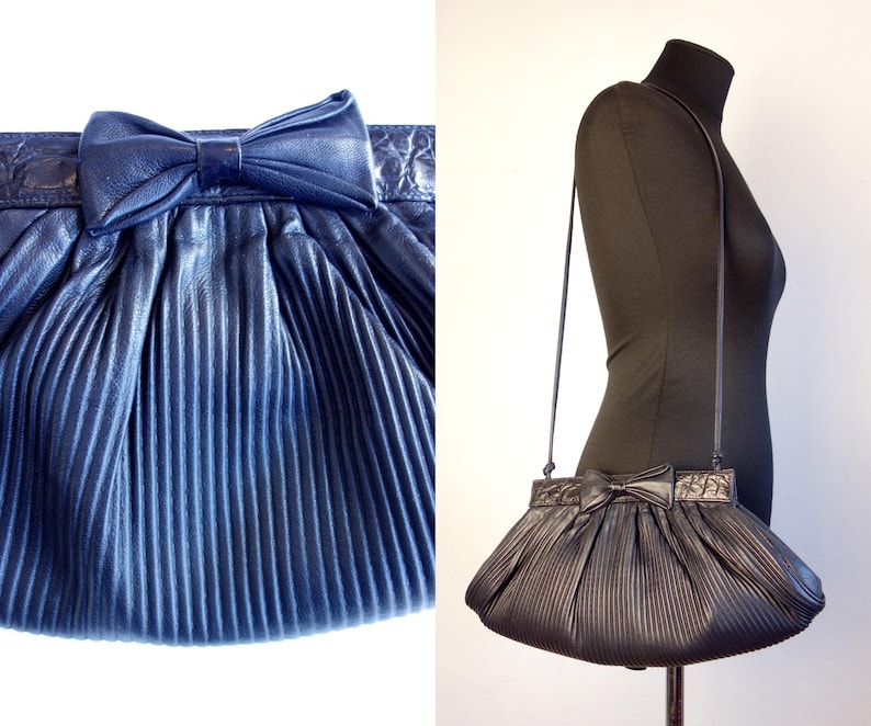 May include: A navy blue pleated handbag with a bow detail. The bag has a long strap and a textured top. The bag is displayed on a mannequin. The bag is a dark blue color.