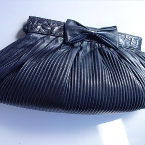 May include: A navy blue pleated clutch with a bow detail. The clutch has a textured, ridged design and a decorative bow on the front. The top of the clutch has a textured, embossed design.