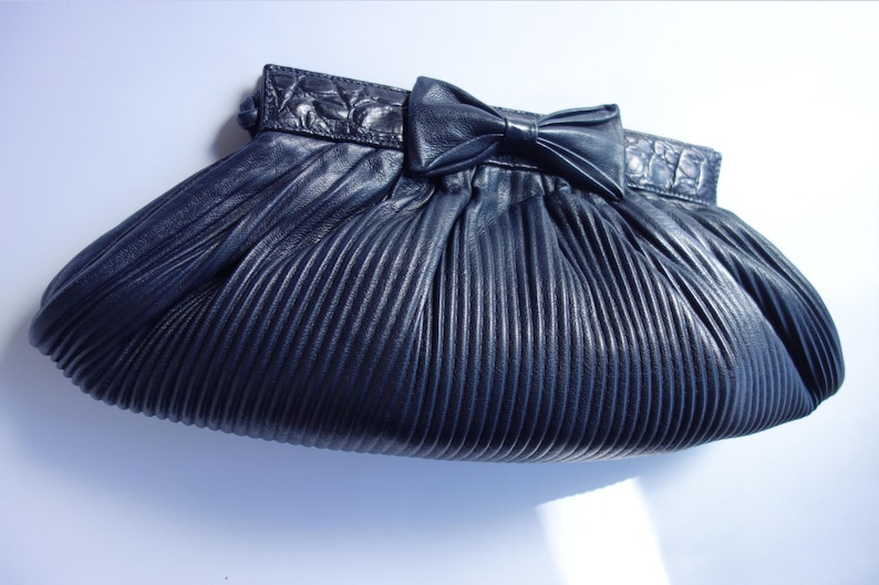May include: A navy blue pleated clutch with a bow detail. The clutch has a textured, ridged design and a decorative bow on the front. The top of the clutch has a textured, embossed design.
