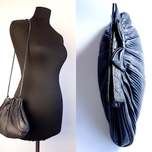 May include: Black leather shoulder bag with a pleated design. The bag has a drawstring closure and a thin shoulder strap. The bag is displayed on a black mannequin. The bag is also shown from a side view.