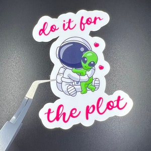 May include: White sticker with a cartoon astronaut hugging a green alien. The sticker says "do it for the plot" in pink lettering.