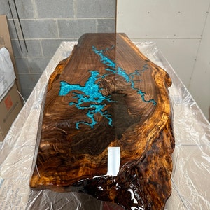 Puede incluir: Una gran tapa de mesa de madera con un borde natural y un relleno de resina epoxi azul turquesa. La resina tiene un patr&oacute;n de remolino y es brillante.