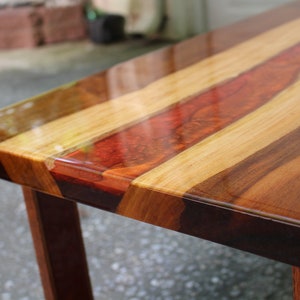 Puede incluir: Primer plano de una mesa de madera con una tapa de resina epoxi transparente. La mesa tiene un acabado de madera natural con una incrustaci&oacute;n de resina epoxi roja, naranja y amarilla. La mesa est&aacute; hecha de diferentes tipos de madera, incluyendo nogal, arce y cerezo.