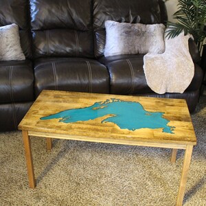 Puede incluir: Una mesa de centro de madera con una incrustaci&oacute;n de resina epoxi azul turquesa en forma de lago. La mesa tiene un acabado de madera natural y cuatro patas.
