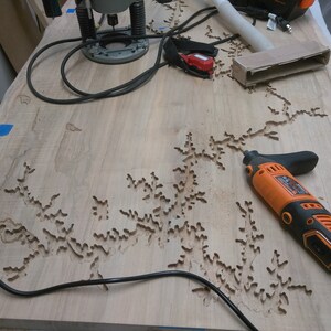 Puede incluir: Primer plano de una superficie de madera con un dise&ntilde;o detallado e intrincado tallado en ella. Una herramienta rotativa naranja y negra est&aacute; sobre la superficie, junto con un cable el&eacute;ctrico negro.