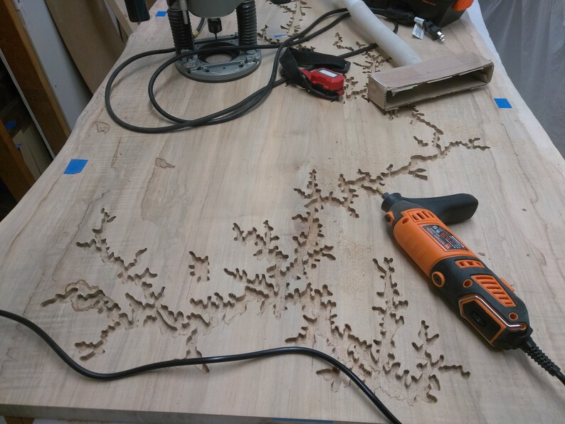Puede incluir: Primer plano de una superficie de madera con un dise&ntilde;o detallado e intrincado tallado en ella. Una herramienta rotativa naranja y negra est&aacute; sobre la superficie, junto con un cable el&eacute;ctrico negro.