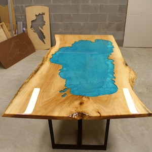 Puede incluir: Una mesa de madera con una tapa de borde vivo y una incrustaci&oacute;n de resina epoxi azul turquesa en forma de lago. La mesa tiene una base de metal negro.