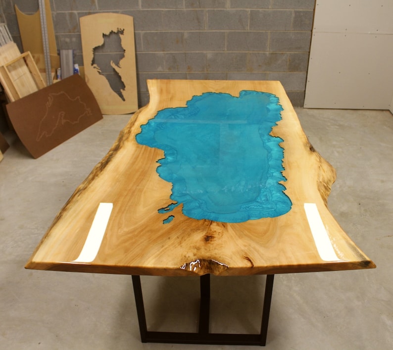 Puede incluir: Una mesa de madera con una tapa de borde vivo y una incrustaci&oacute;n de resina epoxi azul turquesa en forma de lago. La mesa tiene una base de metal negro.