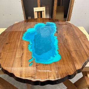 Puede incluir: Una mesa redonda de madera con un borde vivo y una incrustaci&oacute;n de resina epoxi azul y verde en forma de lago.