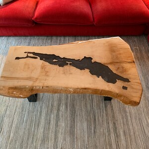 Puede incluir: Una mesa de centro de madera natural con un dise&ntilde;o de borde vivo. La superficie de la mesa tiene un r&iacute;o marr&oacute;n oscuro que la atraviesa. La mesa est&aacute; sostenida por dos patas de metal negro.