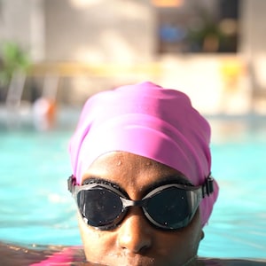 May include: A person wearing a pink swim cap and black swimming goggles is in a swimming pool. The person's face is visible, and the water is blue.