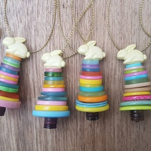 May include: Seven colourful button Christmas tree decorations with a white bunny on top of each tree. The ornaments are hanging from gold chains.