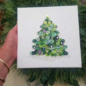 Peut inclure: Une toile blanche avec un sapin de No&euml;l en boutons verts. L'arbre est d&eacute;cor&eacute; de boutons verts, violets et blancs, ainsi que de strass argent&eacute;s et roses. L'arbre est entour&eacute; de paillettes blanches, qui ressemblent &agrave; de la neige.