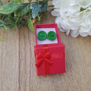 May include: A pair of green button earrings in a red gift box with a red ribbon bow.