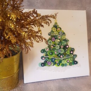 Peut inclure: Une toile blanche avec un sapin de No&euml;l en boutons verts. L'arbre est d&eacute;cor&eacute; de boutons verts, roses et blancs, ainsi que de strass. Le fond est blanc avec quelques points blancs.